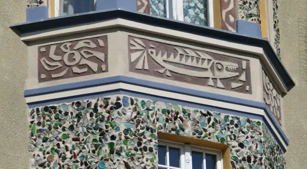 Sgraffito z rybą i abstrakcyjnymi kształtami na fasadzie budynku, ozdobionej mozaiką z kolorowego szkła.