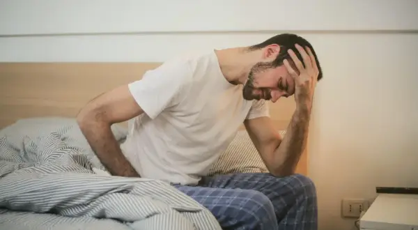 Mann sitzt im Bett, hält sich den Kopf. Mögliche Bewegungsmangel Symptome wie Kopfschmerzen und Müdigkeit sind zu erkennen.