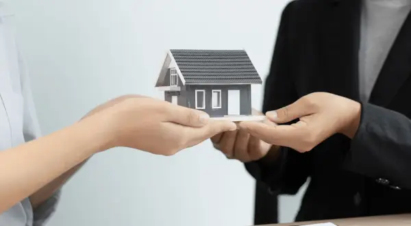 Manos intercambian una maqueta de casa, simbolizando la gestión de alquileres y la transferencia de propiedades.
