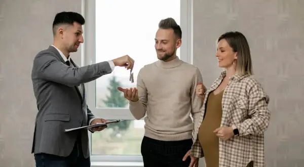 Agente inmobiliario realiza la entrega de llaves de alquiler a una pareja joven, uno de ellos embarazado, en un nuevo hogar.