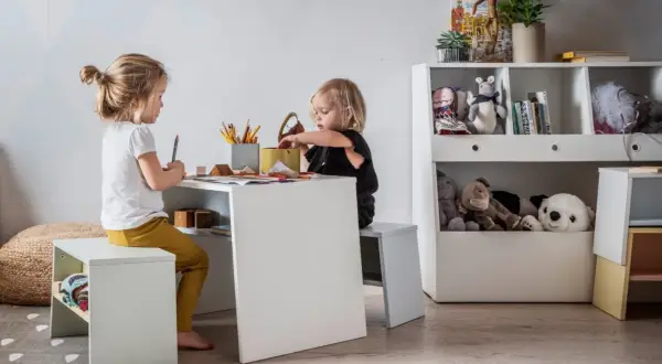 Uroczy kącik dla dziecka w salonie. Dwie dziewczynki rysują przy białym stoliku, otoczone pluszakami i książkami.