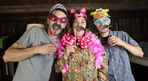 Tres amigos se divierten con disfraces hechos con cosas de casa: un hombre con tiara y bigote, una mujer con orejas de gato y un hombre con máscara de vaca.