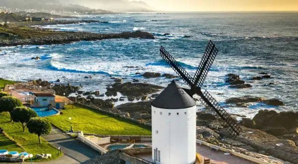 Un muiño do vento blanco con aspas negras se alza junto a un mar azul y rocoso. El paisaje costero es agreste y hermoso.