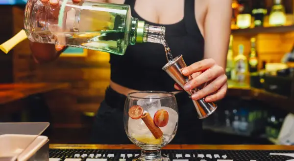 Barback nalewa alkohol z butelki do jiggera, przygotowując drinka z lodem, winogronem i cynamonem.