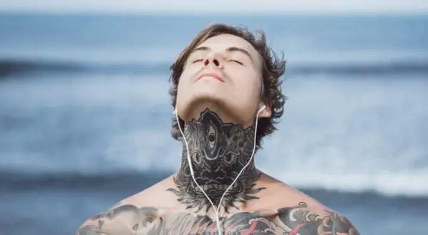 Mężczyzna z tatuażem na szyi i klatce piersiowej, słuchający muzyki na plaży. Jego oczy są zamknięte, a głowa lekko uniesiona.