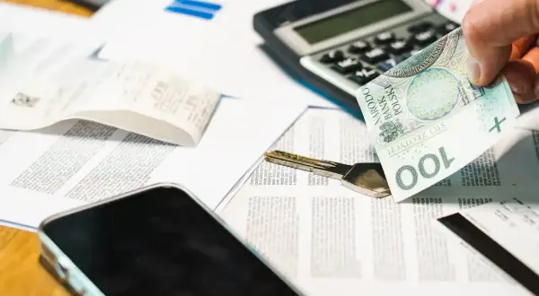 Dłoń trzyma banknot 100 zł nad dokumentami, kluczem i telefonem. To potwierdzenie odbioru gotówki.