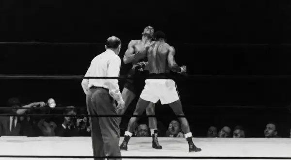 A referee watches as two boxers battle in the ring. One boxer, with his head thrown back, is hit. This intense moment captures the fight for who is the king of boxing.