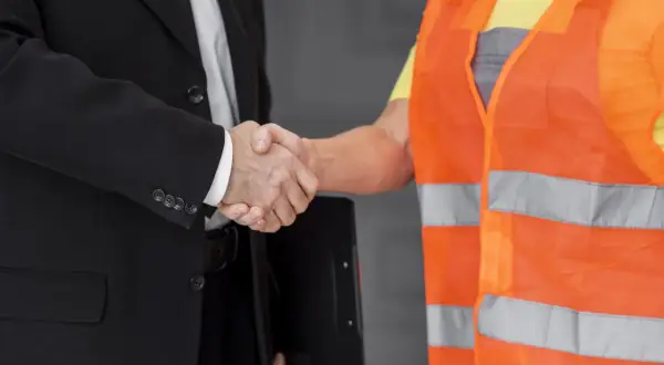 Ein Geschäftsmann im Anzug und ein Arbeiter in Warnweste schütteln sich die Hände. Sie sind Partner des Staates beim Arbeitsschutz.