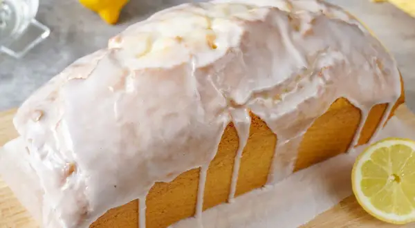 Saftiger zitronenkuchen in Kastenform, überzogen mit süßem Zuckerguss. Ein Zitronenstück liegt daneben.