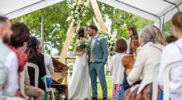 Un couple s'embrasse lors de leur baptême civil, entouré de leurs proches sous une tente décorée de fleurs.