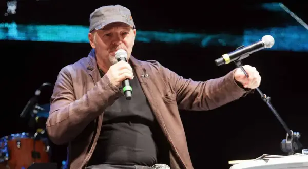 Uomo con cappellino e giacca di pelle marrone parla al microfono, sembra che sono ancora in coma, ma è sul palco.