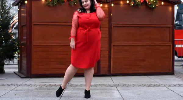 Kobieta w czerwonej sukience prezentuje eleganckie komplety damskie ze spodniami XXL, pozując na tle świątecznych straganów.