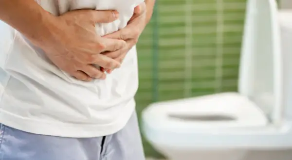 Mann hält sich den Bauch, deutet auf mögliche Harnverhalt Symptome. Im Hintergrund eine Toilette.