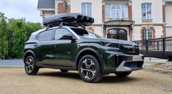 Nowy C3 Aircross w ciemnozielonym kolorze z czarnym boxem dachowym przed eleganckim budynkiem.