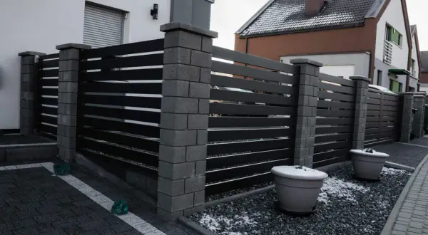 Nowoczesne ogrodzenie z czarnych paneli i kamiennych słupków. Jego wysokość zapewnia prywatność. Dwa duże donice z zimowym akcentem śniegu.