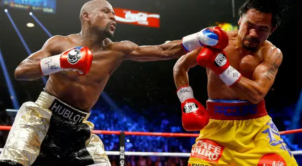 Two boxers in a ring, one throwing a punch. The inside fighter, with gold shorts, connects with his opponent.