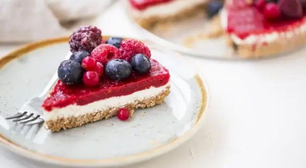 Una fetta di cheesecake ai frutti di bosco, con una glassa rossa brillante e bacche fresche, su un piatto con una forchetta.