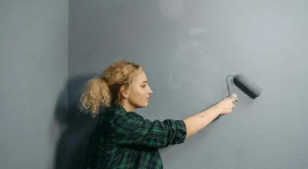 Mujer rubia con coleta pinta pared gris con rodillo. Se pregunta cuanto cuesta pintar un piso.