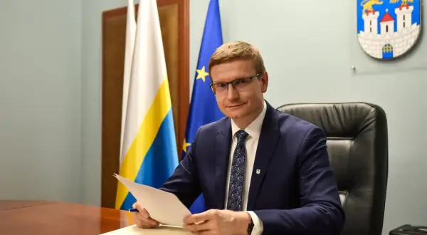 Mężczyzna w garniturze siedzi przy biurku, trzymając dokumenty. Za nim flagi i herb miasta, symbolizujące jego rolę w obszarze prawo administracyjne.