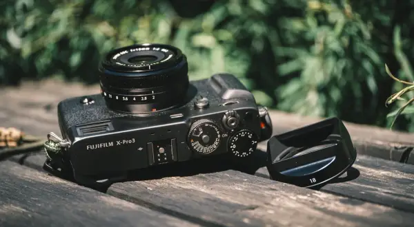 A Fujifilm X-Pro3 camera with a lens and lens hood rests on a wooden surface, ready for a photography review.