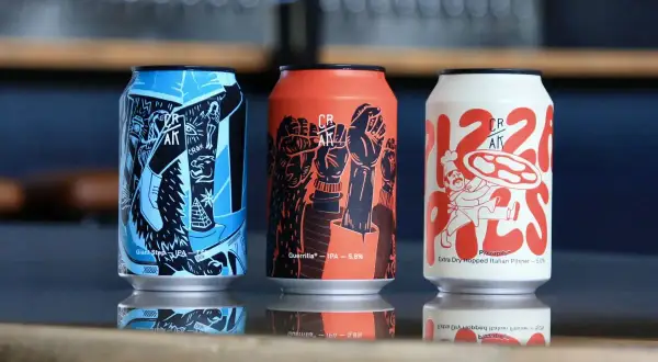 Tre lattine di crak beer con grafiche artistiche: una blu con piramide, una arancione con pugni alzati, una bianca con un pizzaiolo.