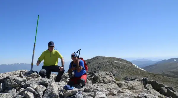 Dos excursionistas celebran en la cima tras una **cuerda larga ruta**. Un poste verde marca el camino.