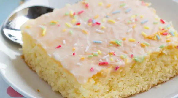 Ein Stück saftiger Fantakuchen vom Blech, mit Zuckerguss und bunten Streuseln bestreut, auf einem Teller mit Löffel.