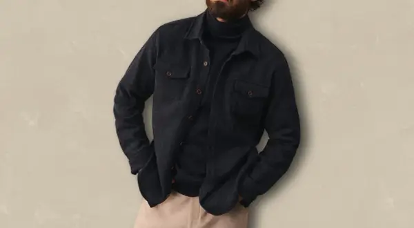Un homme porte une surchemise foncée ouverte sur un col roulé bleu marine, avec un pantalon beige. Idéal pour savoir comment porter une surchemise avec style.