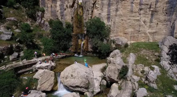 Un hermoso salto de agua cae en una poza natural rodeada de rocas y vegetación. Varias personas disfrutan del paisaje y del agua.