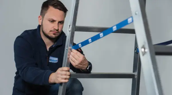 Mężczyzna w granatowej bluzie z logo KRAUSE przeprowadza badanie drabin, sprawdzając mocowanie niebieskiej taśmy zabezpieczającej.