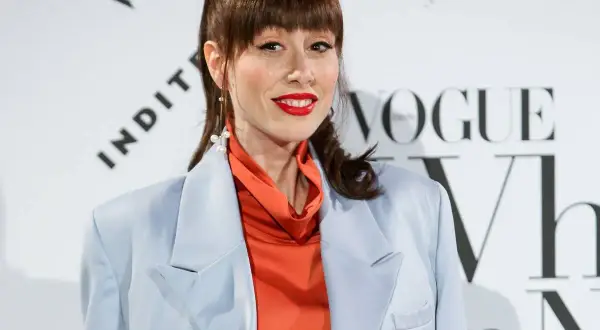 Natalia Ferviu, con su coleta alta y labios rojos, luce un traje azul pálido y un top naranja vibrante.