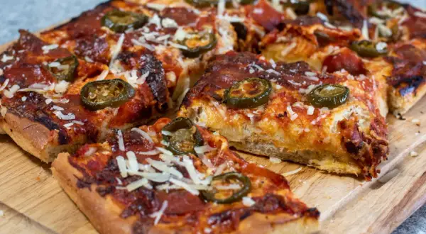 En saftig pan pizza med pepperoni och jalapeños, toppad med riven ost, ligger på en träskärbräda.