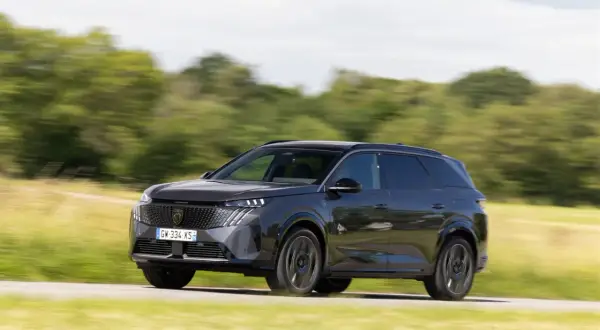 Ciemnoszary Peugeot 5008 pędzi po drodze, otoczony zielenią pól i drzew.