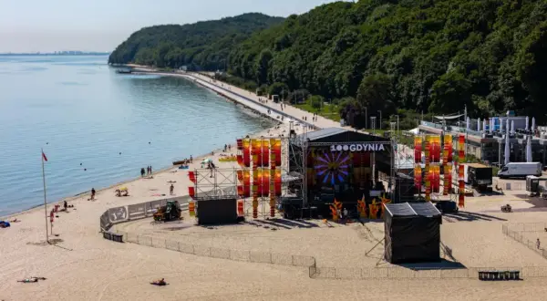 Scena festiwalowa na plaży w Gdyni, z widokiem na morze i las. Kolorowe dekoracje i logo "100 Gdynia" na scenie.