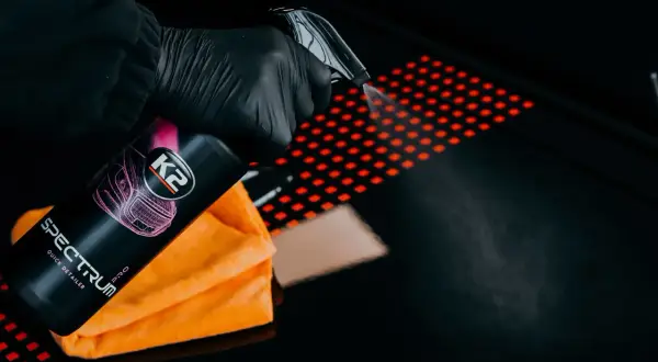 Ręka w czarnej rękawiczce rozpyla quick detailer K2 Spectrum Pro na czarną powierzchnię z czerwonym wzorem. Pomarańczowa mikrofibra obok.