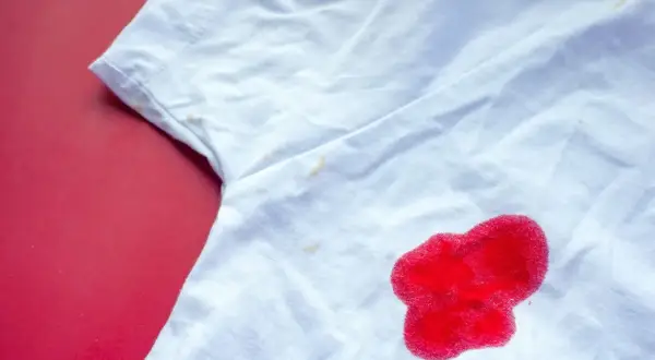 Biała koszulka z plamą z czerwonego płynu i kilkoma mniejszymi, żółtymi plamkami.