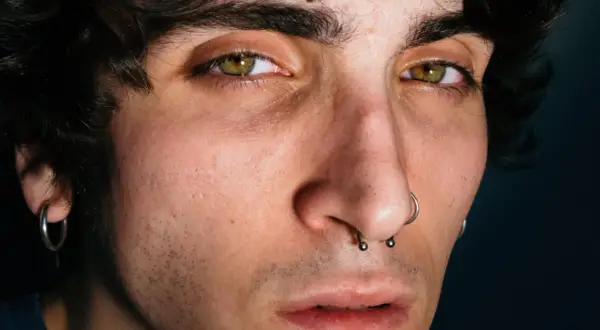 Primo piano di un giovane con occhi verdi penetranti, capelli scuri e piercing al naso. Giuse the Lizia, un volto intenso.
