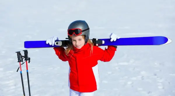 Uśmiechnięta dziewczynka w czerwonej kurtce i białych spodniach niesie narty na ramionach. Gotowa na ferie zimowe 2026 lubuskie.