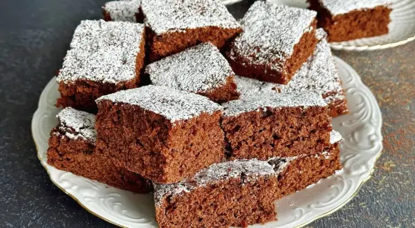 Pyszne ciasto czekoladowe pokrojone w kostkę, posypane cukrem pudrem, ułożone na białym talerzu.