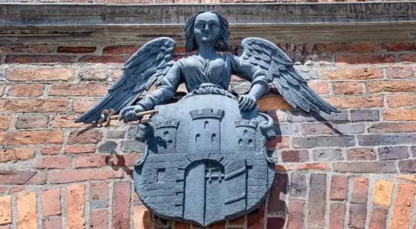 Kamienna rzeźba anioła z kluczem i tarczą z zamkiem, symbolizująca herb Torunia, zdobi ceglaną ścianę.