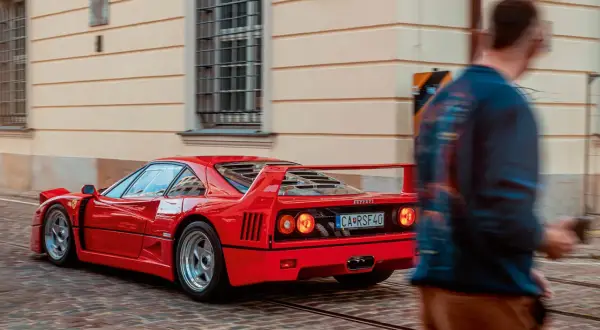 Czerwone Ferrari F40 z charakterystycznym spojlerem i okrągłymi światłami mija brukowaną ulicę, a obok przechodzi rozmyta postać mężczyzny.