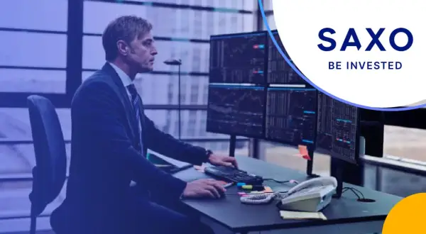 Mężczyzna w garniturze analizuje wykresy na monitorach w biurze Saxo Bank Polska. "Be invested" - hasło zachęca do inwestowania.