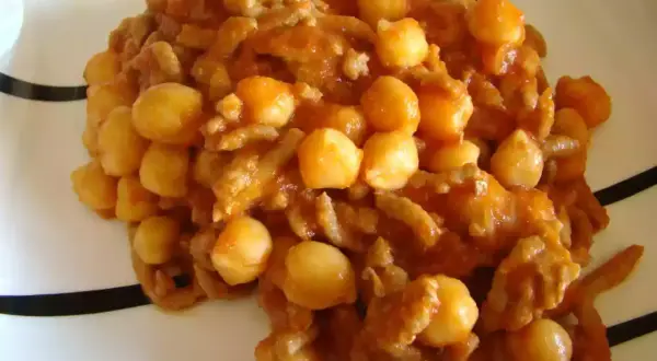 Un plato de garbanzos con carne picada, cocinado en una salsa rojiza y servido en una vajilla blanca con rayas negras.
