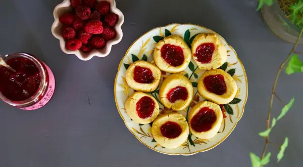 Hallongrottor recept: småkakor med hallonsylt på en vacker tallrik, bredvid en skål med färska hallon och en burk sylt.