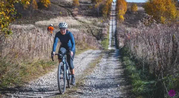 Kobieta na rowerze pokonuje kamienistą ścieżkę w Dolinkach Krakowskich. Jesienne drzewa i trawy tworzą malowniczy krajobraz.