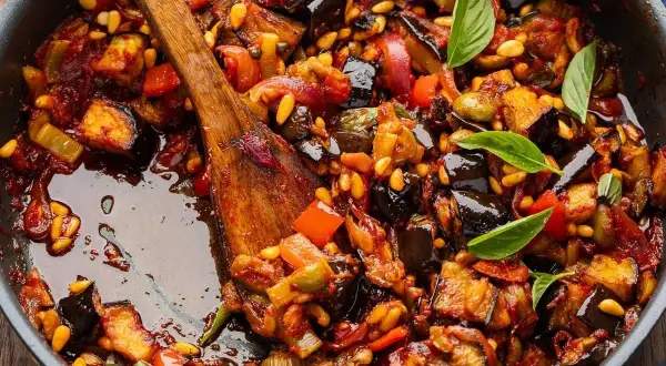 En varm, mustig siciliansk caponata med aubergine, paprika och pinjenötter i en panna, redo att serveras.
