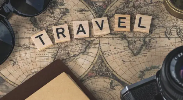 Drewniane klocki z napisem TRAVEL na starej mapie świata, obok okulary, aparat i kompas.