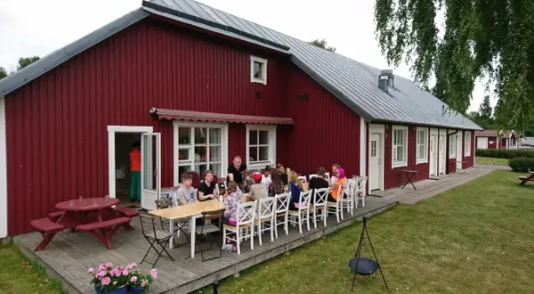Gästerna njuter av en måltid utomhus vid Sjötorp vandrarhem. En grupp vänner samlas runt borden på altanen framför den röda byggnaden.