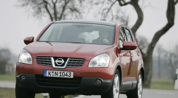 Czerwony Nissan Qashqai 1.6 benzyna, często chwalony w opiniach, jedzie po drodze wśród drzew.