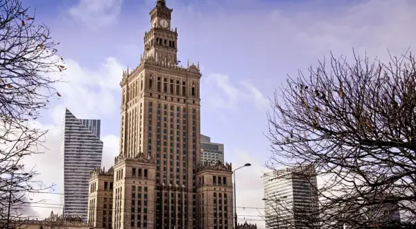 Kulturpalatset Warszawa reser sig stolt mot en molnig himmel, omgiven av moderna skyskrapor och kala trädgrenar.